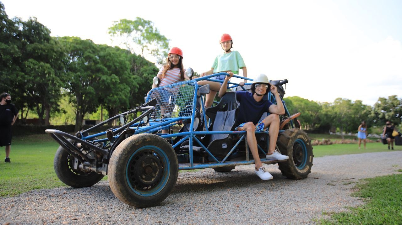 Três pessoas, usando capacetes, sentam-se em um buggy azul em uma estrada de terra. Uma pessoa está sentada no teto do buggy, sorrindo.