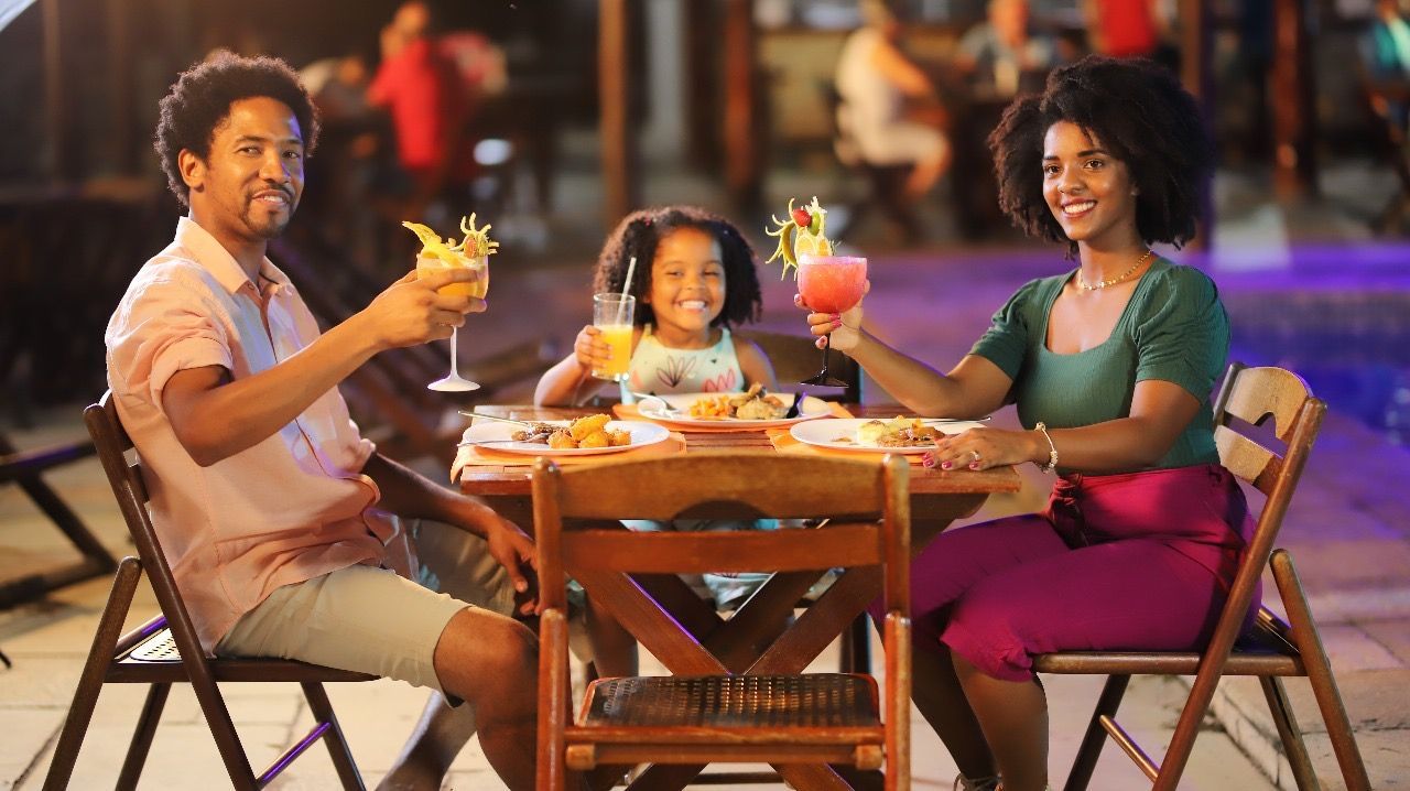 Família brindando com bebidas em um restaurante ao ar livre. Pai de rosa, mãe de verde e roxo, e filho em um vestido florido.