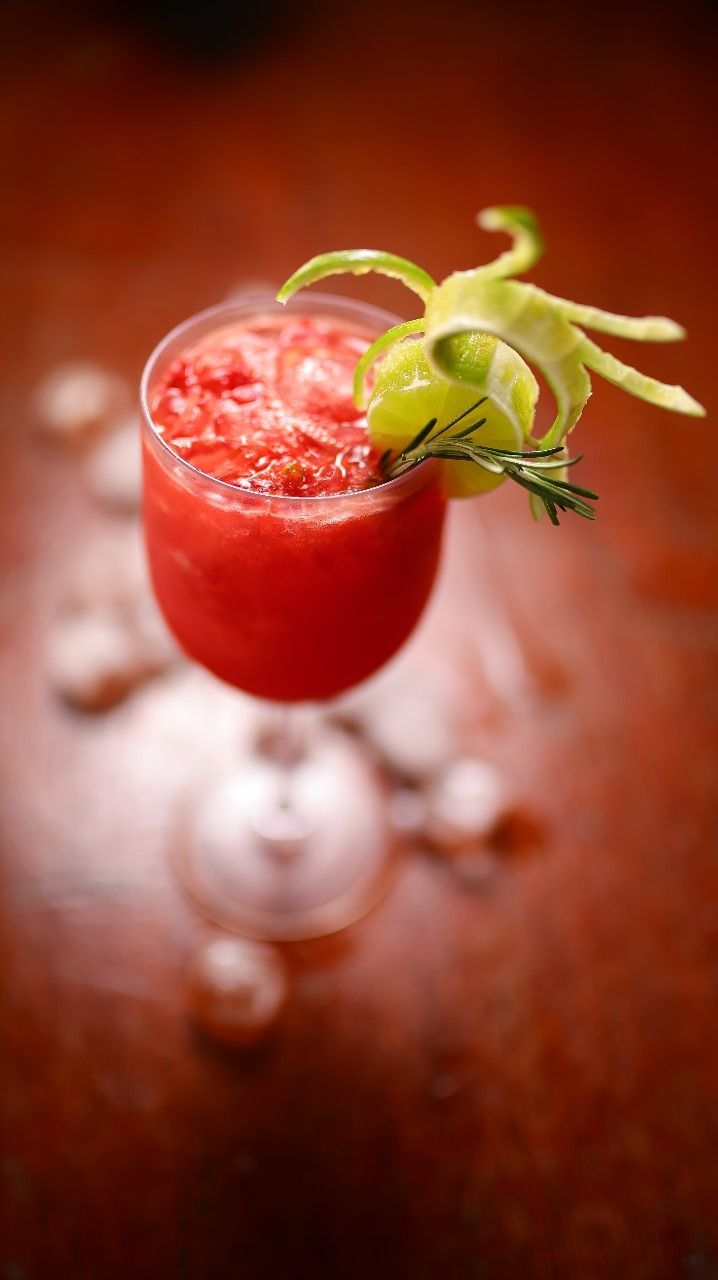 Um coquetel vermelho vibrante em uma taça de haste, decorado com uma rodela de limão verde e alecrim, sobre uma superfície marrom com gelo.
