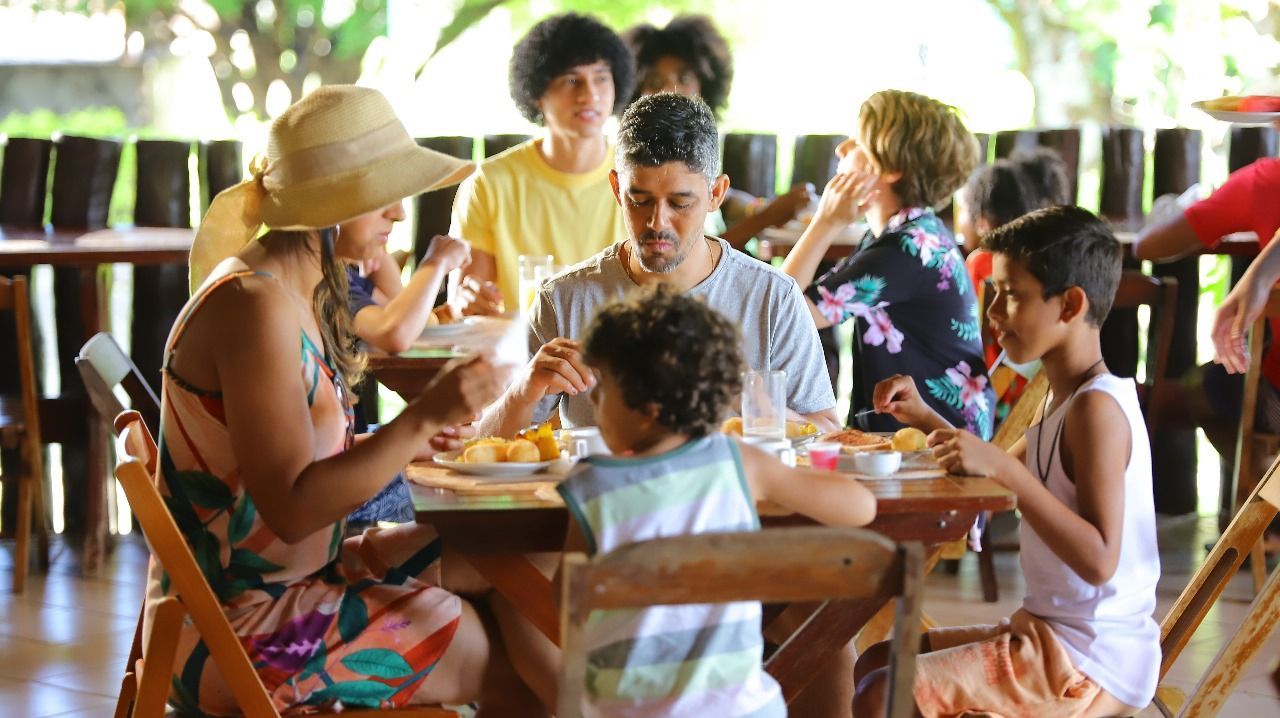 Uma família e outras pessoas jantando em mesas de madeira em um restaurante ao ar livre. As pessoas sorriem e conversam.