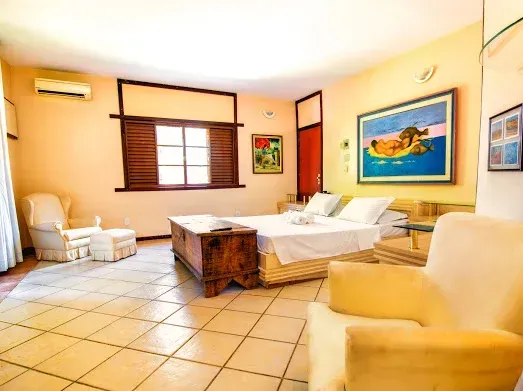 Quarto bem iluminado com cama, área de estar e obras de arte. Possui cômoda de madeira, piso de cerâmica branca e paredes amarelas.