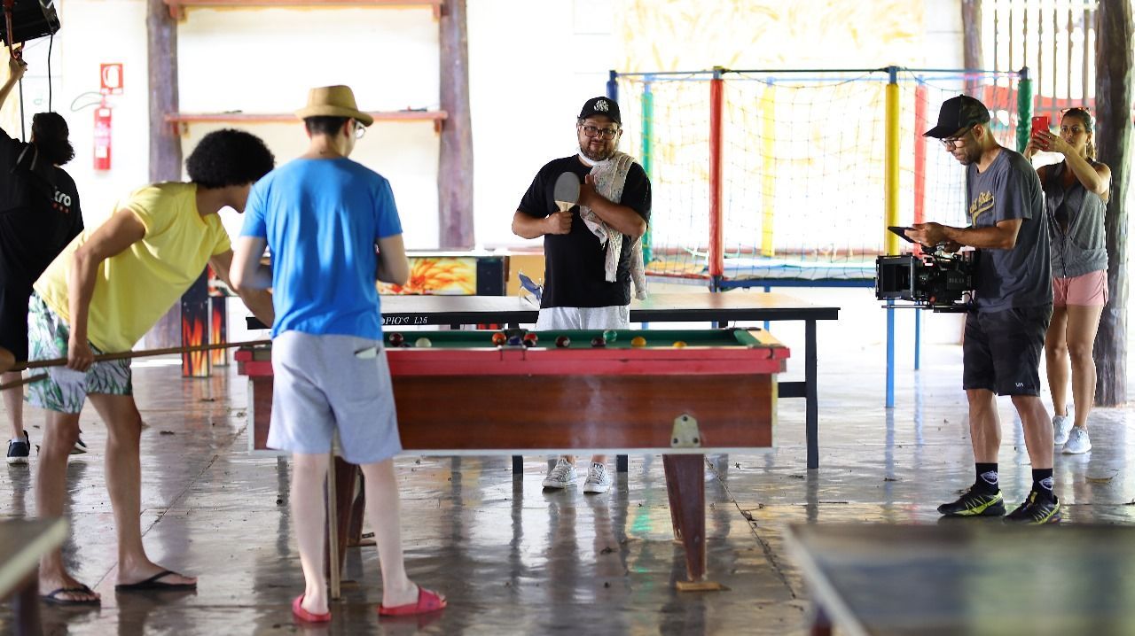 Pessoas jogando pebolim em ambientes fechados. Outras assistem, uma delas segurando um cachorrinho.