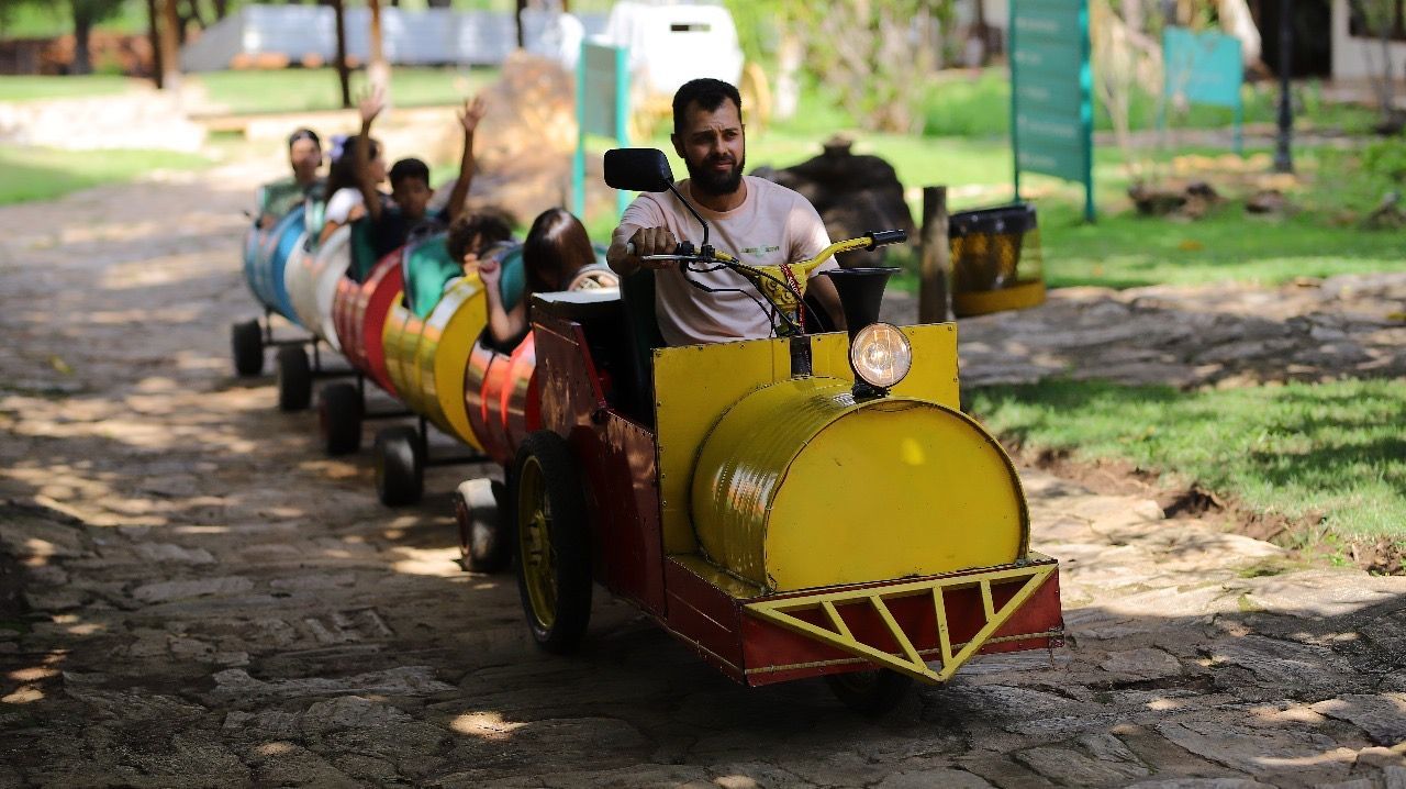 Um trem colorido, feito em casa, com passageiros, percorre um caminho arenoso ao ar livre. Um homem dirige o vagão da frente.