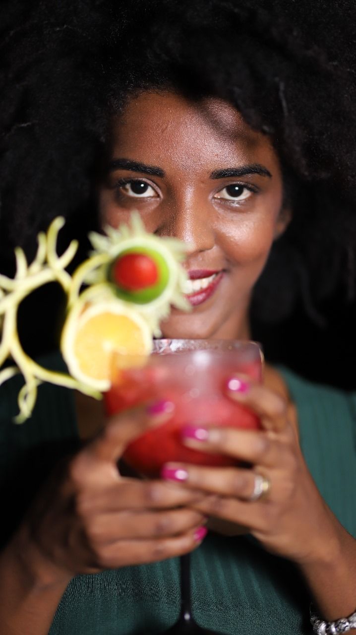 Mulher de cabelos escuros sorrindo, segurando um coquetel vermelho decorado com fatias de pepino, limão e lima.