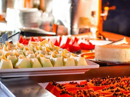Bandejas com diversas frutas cortadas, incluindo melão e mamão, em um bufê. Um pequeno cheesecake ao fundo.