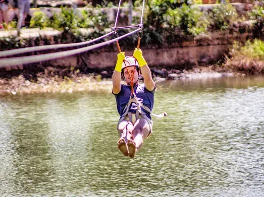 Pessoa em uma tirolesa sobre a água, usando capacete e luvas, com os braços levantados e uma expressão animada.