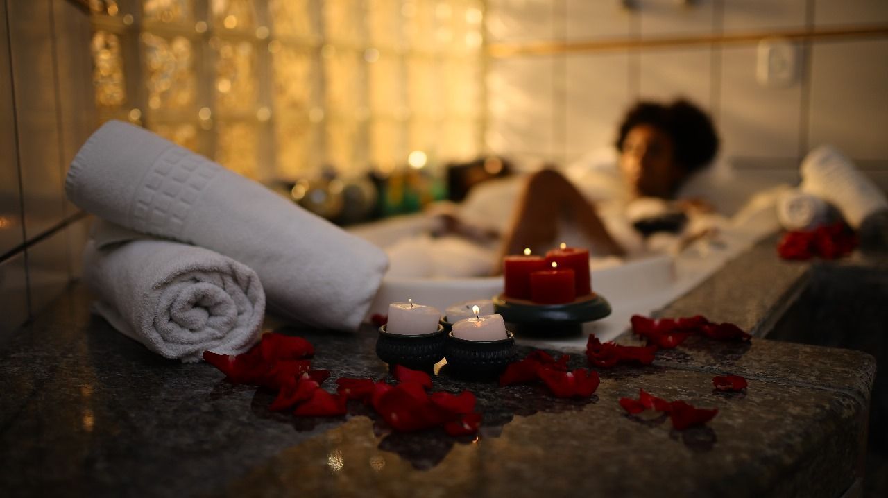 Toalhas brancas enroladas, velas e pétalas de rosa sobre uma superfície de mármore; uma pessoa relaxa em uma banheira em um banheiro mal iluminado.