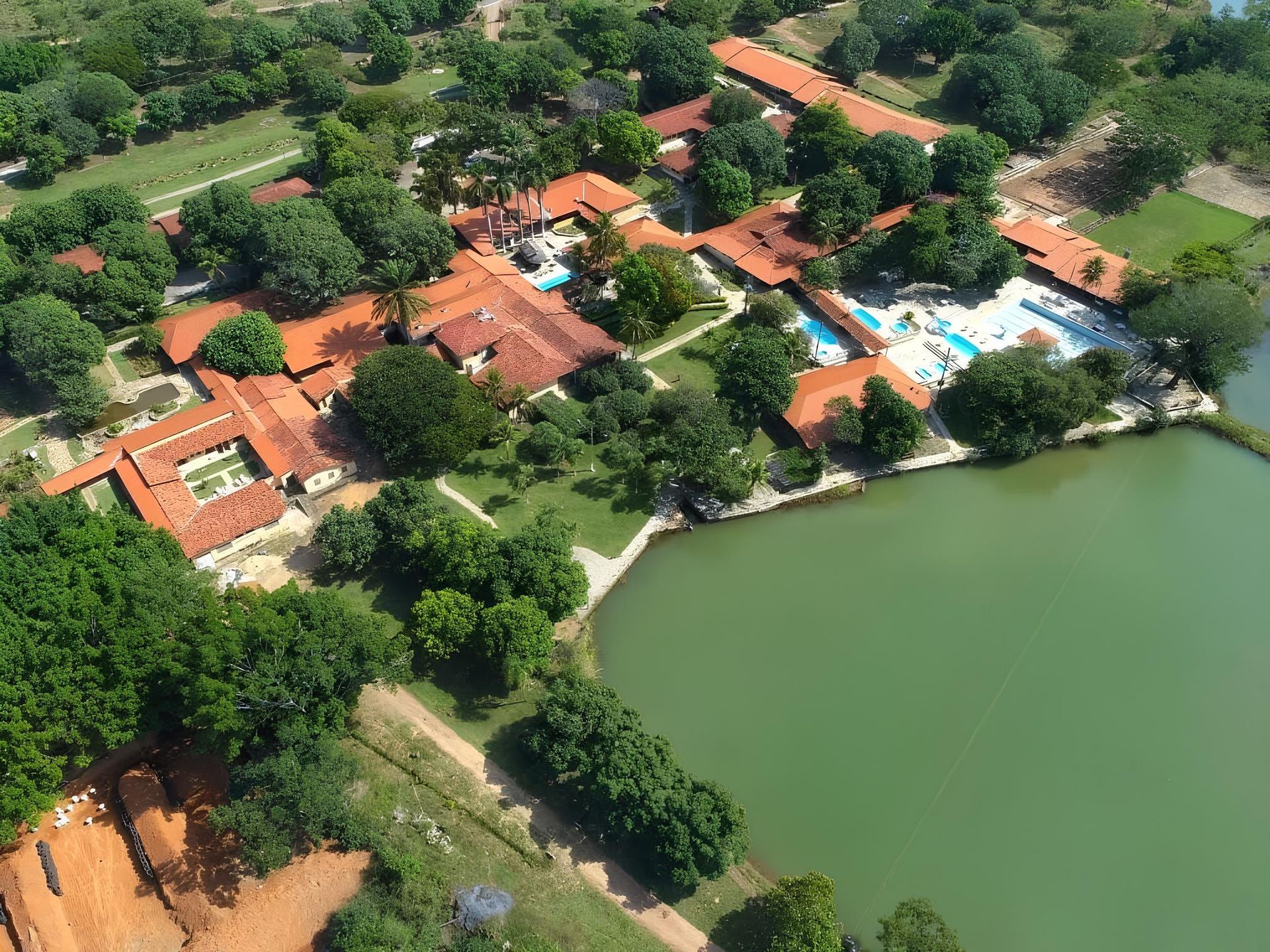 Vista aérea de um resort com telhados vermelhos, piscinas e um lago cercado por árvores verdes.