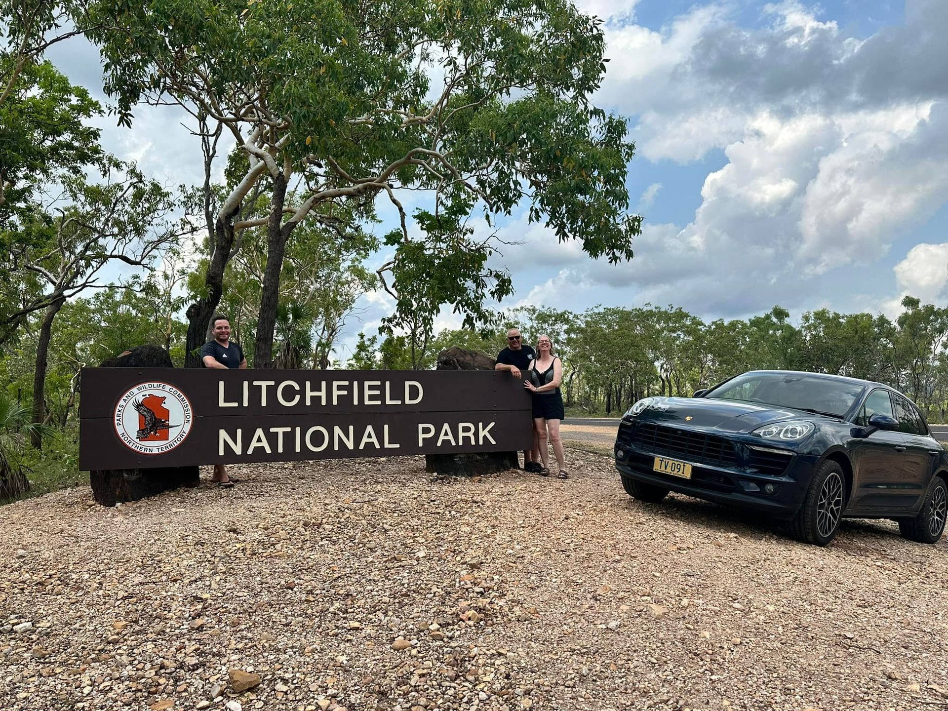Three people pose near a Litchfield National Park sign and a blue SUV on a gravel road under a cloudy sky — Litchfield VIP Tours In Darwin City, NT