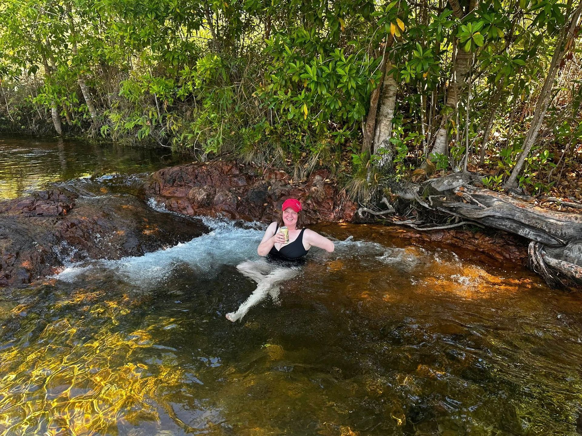 Woman in Black Swimsuit and Red Hat Sits in Clear, Shallow Water by Rocks and Trees — Litchfield VIP Tours In Darwin City, NT