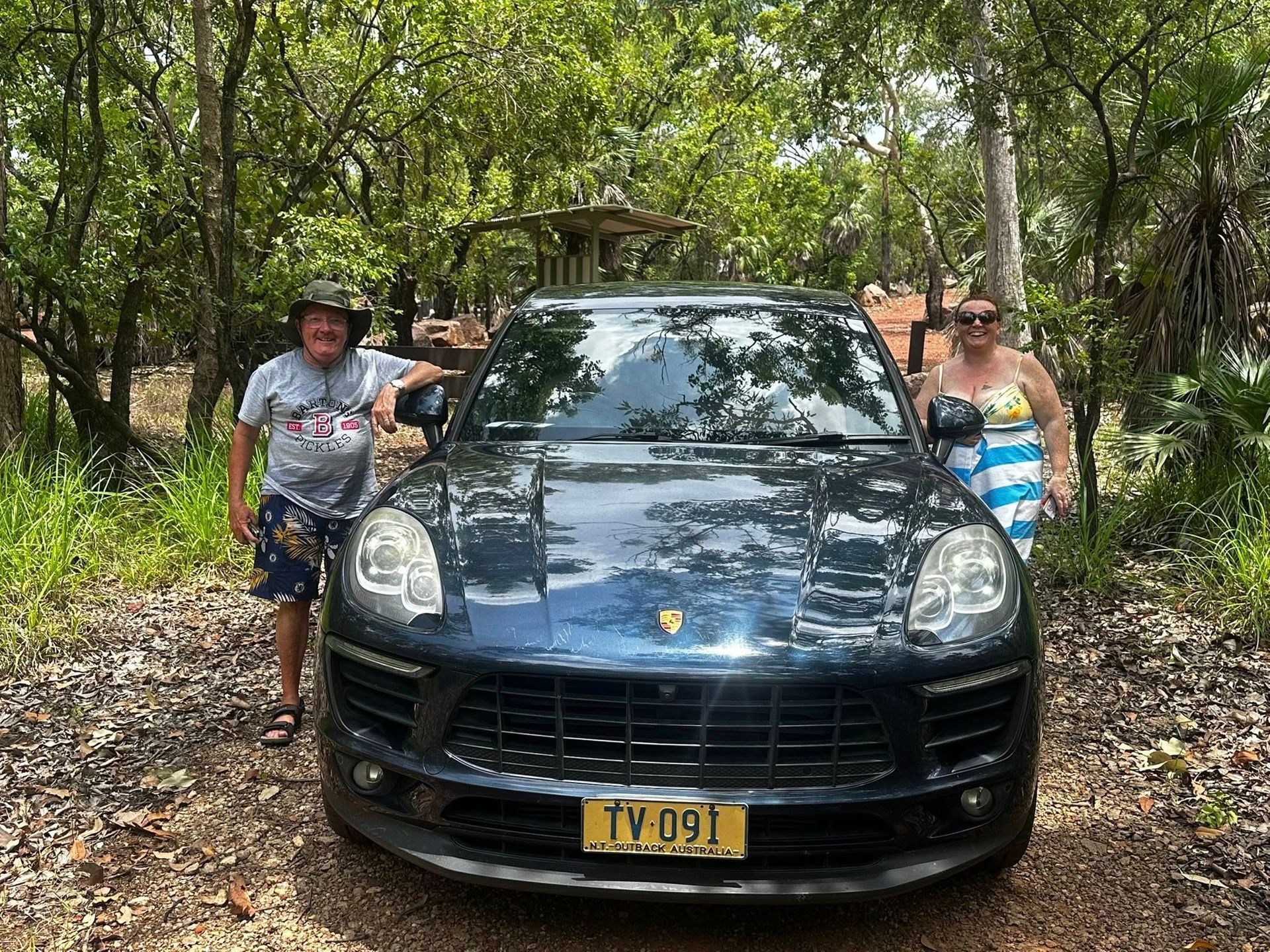 Couple posing with a dark blue Porsche SUV on a dirt road, trees in background — Litchfield VIP Tours In Darwin City, NT