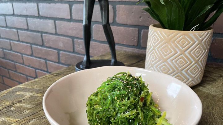 A bowl of green vegetables is sitting on a wooden table next to a potted plant.