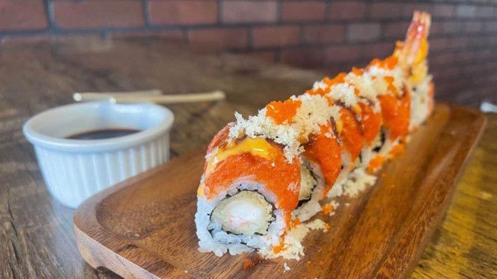 A sushi roll is sitting on a wooden cutting board next to a bowl of dipping sauce.