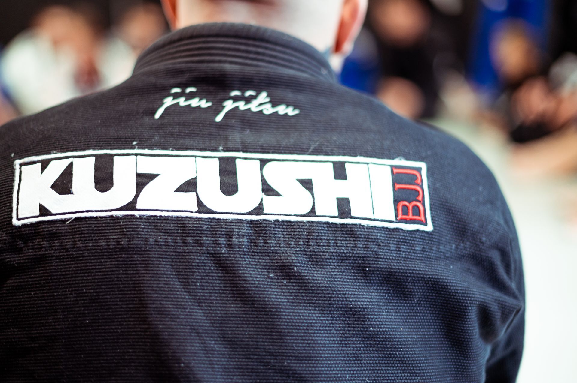 Eine Person in einem blauen Gi beobachtet zwei andere beim Ringen auf der Matte in einem hell erleuchteten Dojo.