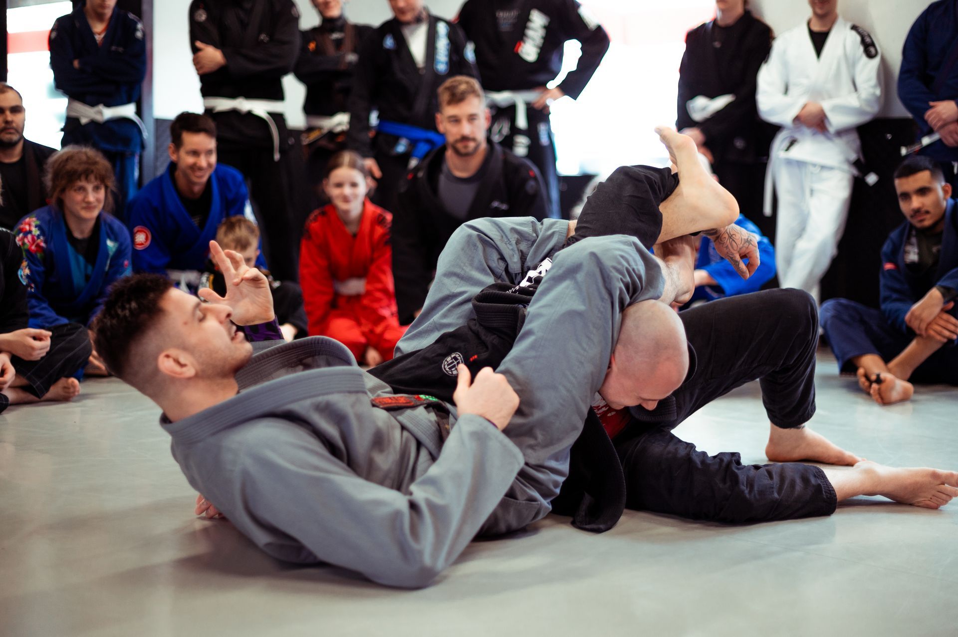 Zwei Personen in Gis trainieren auf dem Boden Brazilian Jiu Jitsu (BJJ), beobachtet von einer Gruppe von Menschen im Hintergrund.
