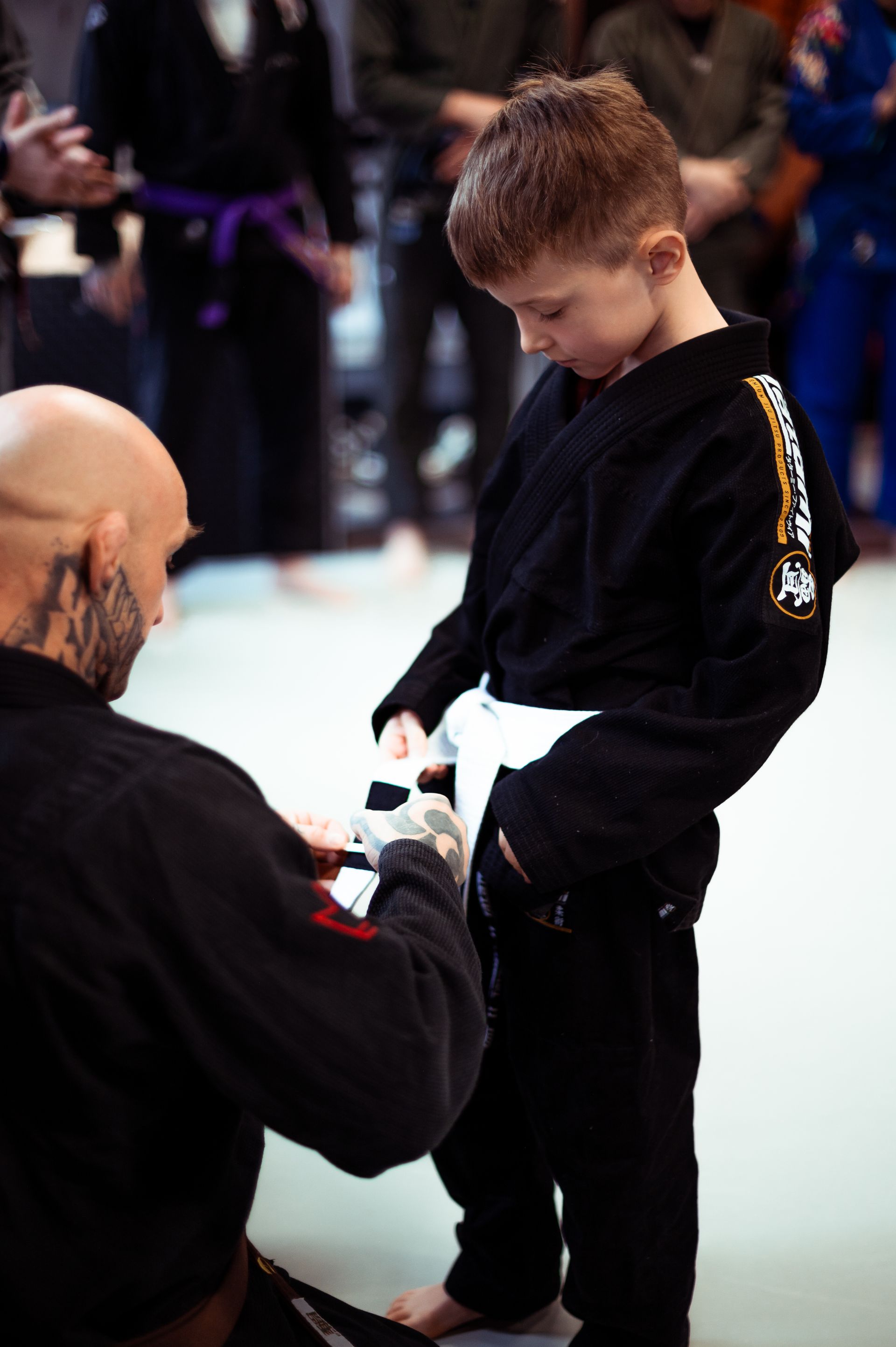 Eine Person in schwarzer Kampfsportuniform richtet in einer Turnhalle den weißen Gürtel eines Schülers in schwarzem Gi.