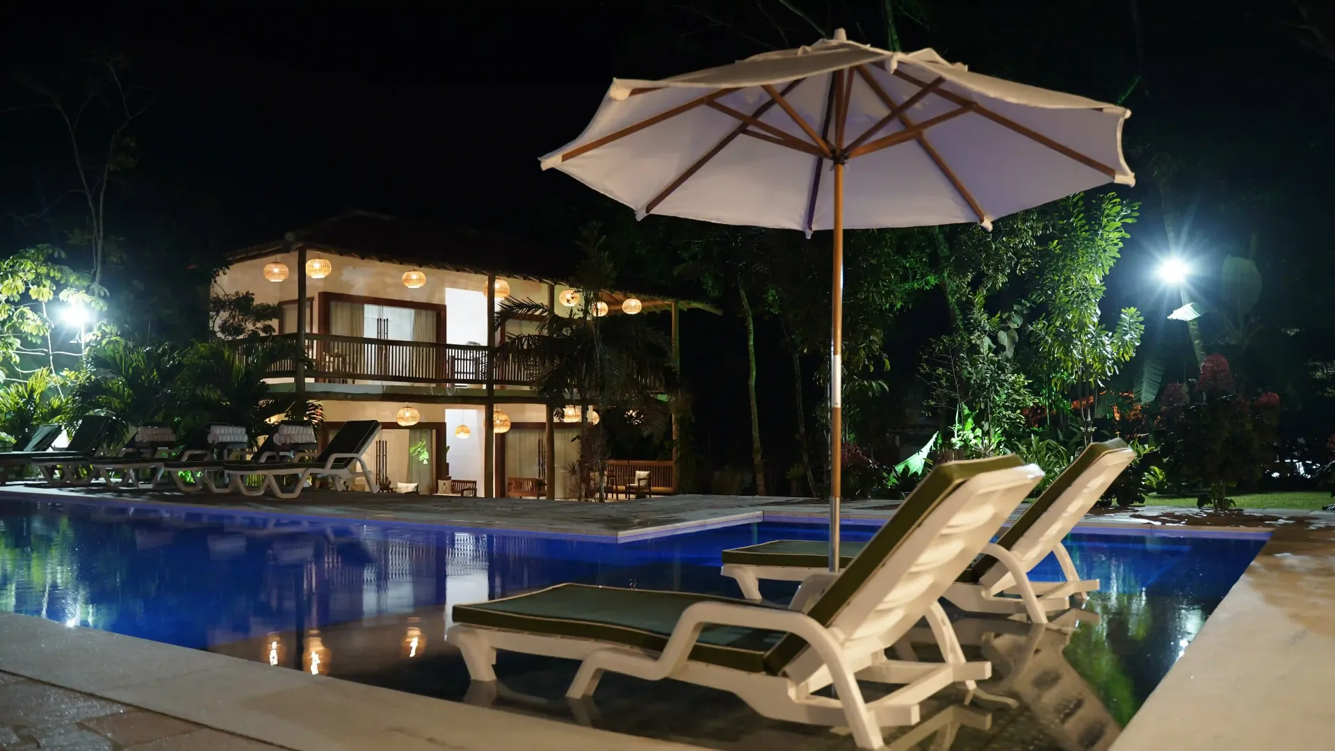 Uma piscina à noite com cadeiras e um guarda-sol - pousada-recanto-caxando