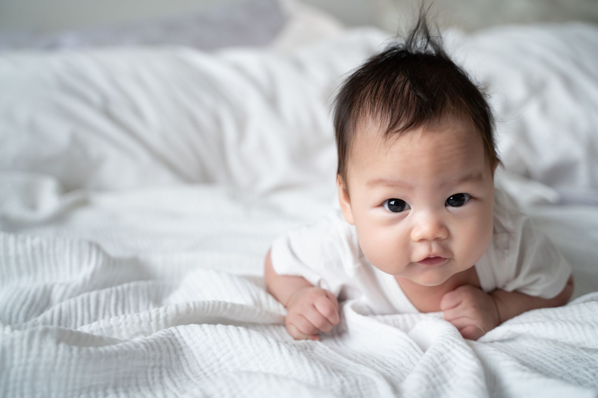 Baby lying on a white bed, looking at the camera. Baby has spiky hair and is wearing white clothes.