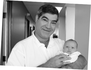 A person in a white collared shirt smiling while holding a newborn baby in a room with a hallway in the background.