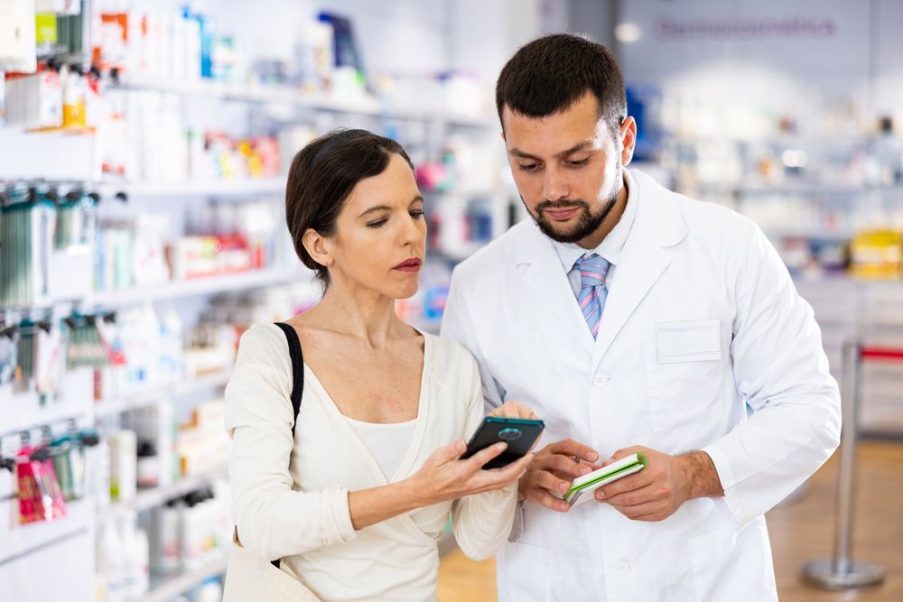 Friendly Man Pharmacist Helping Young Adult Woman WithElectronic Prescription In Her Phone Choosing Medical Supplies At Drugstore — Scripts on File & E-Scripts in Wulguru, QLD