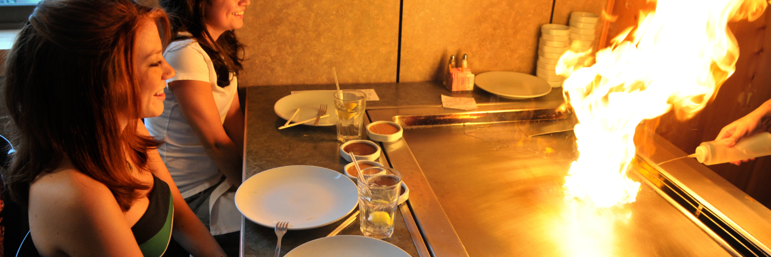 Two women are sitting at a table in front of a stove with flames coming out of it.