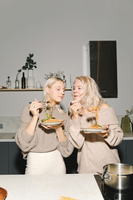 Two women are eating spaghetti together in a kitchen.