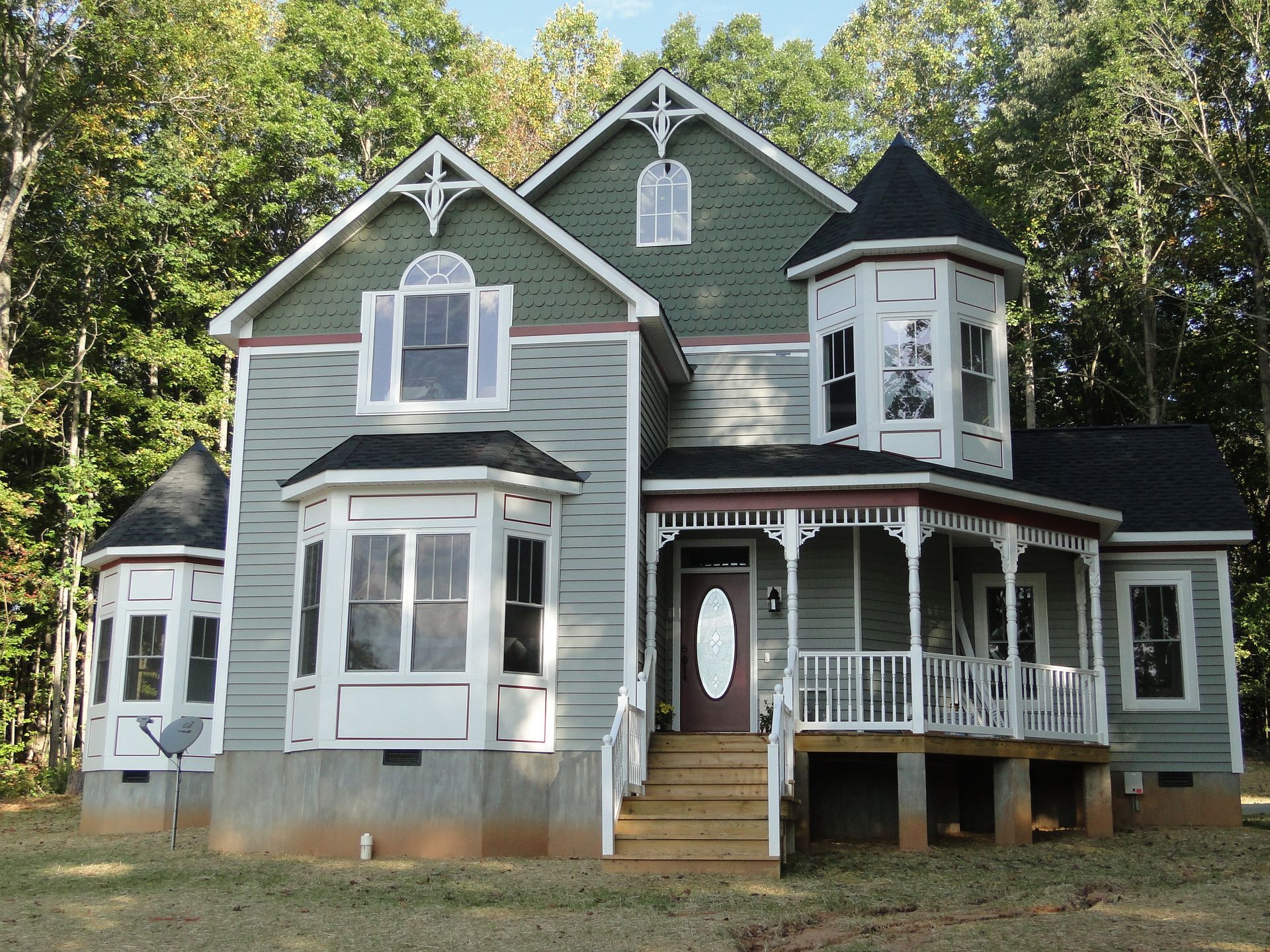 A multi-story Victorian-style house with green and gray siding, a wraparound porch, and a turret, surrounded by trees.