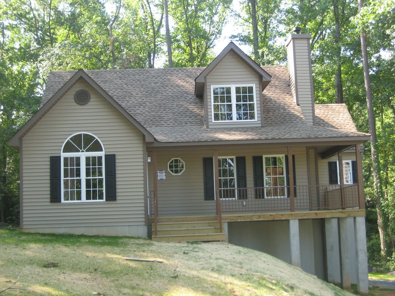 A tan, two-story house with a wooden porch, arched window, and chimney, nestled in a wooded, sloped landscape.