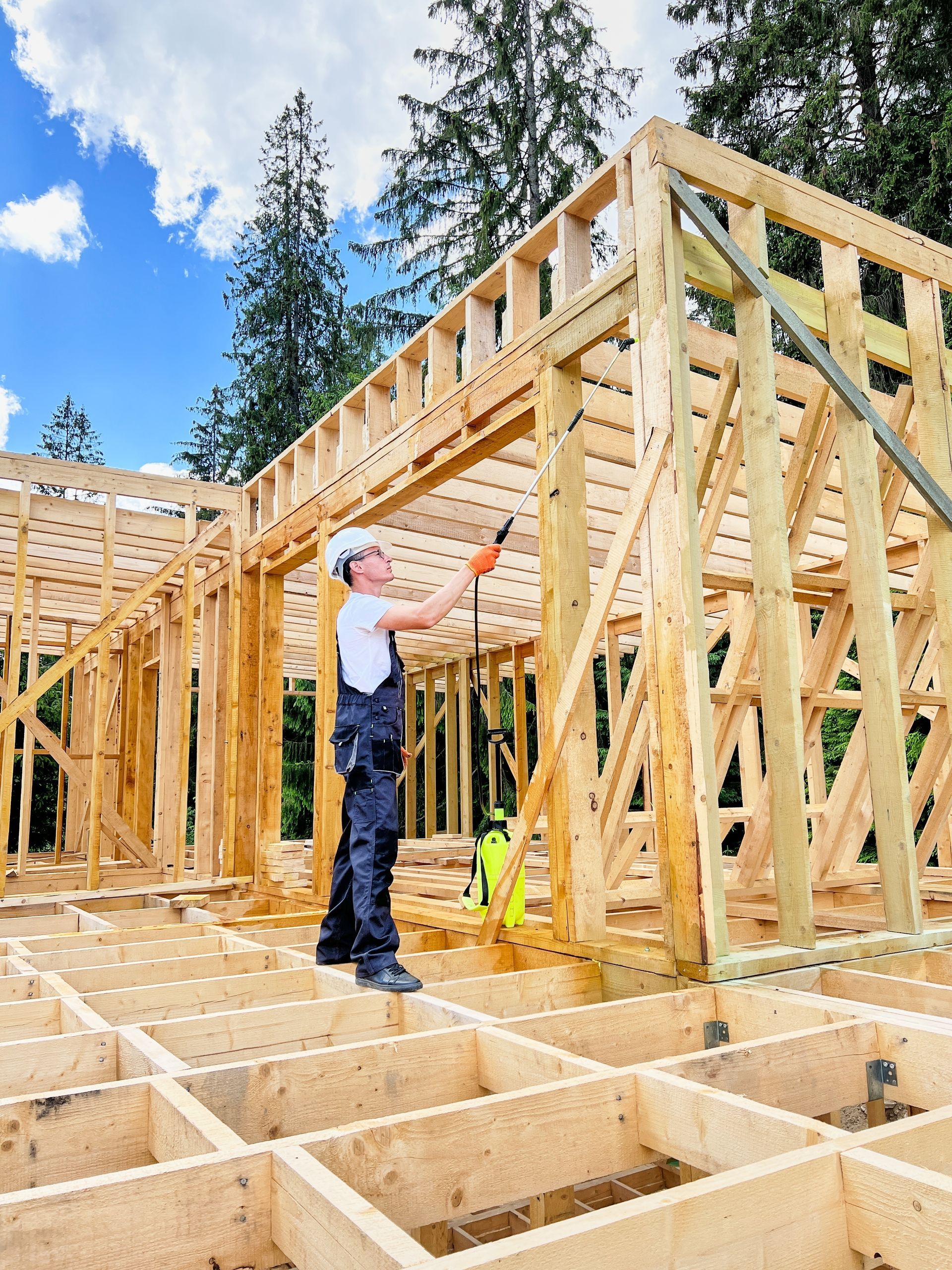 Construction worker in overalls works on a wooden house frame under a blue sky.