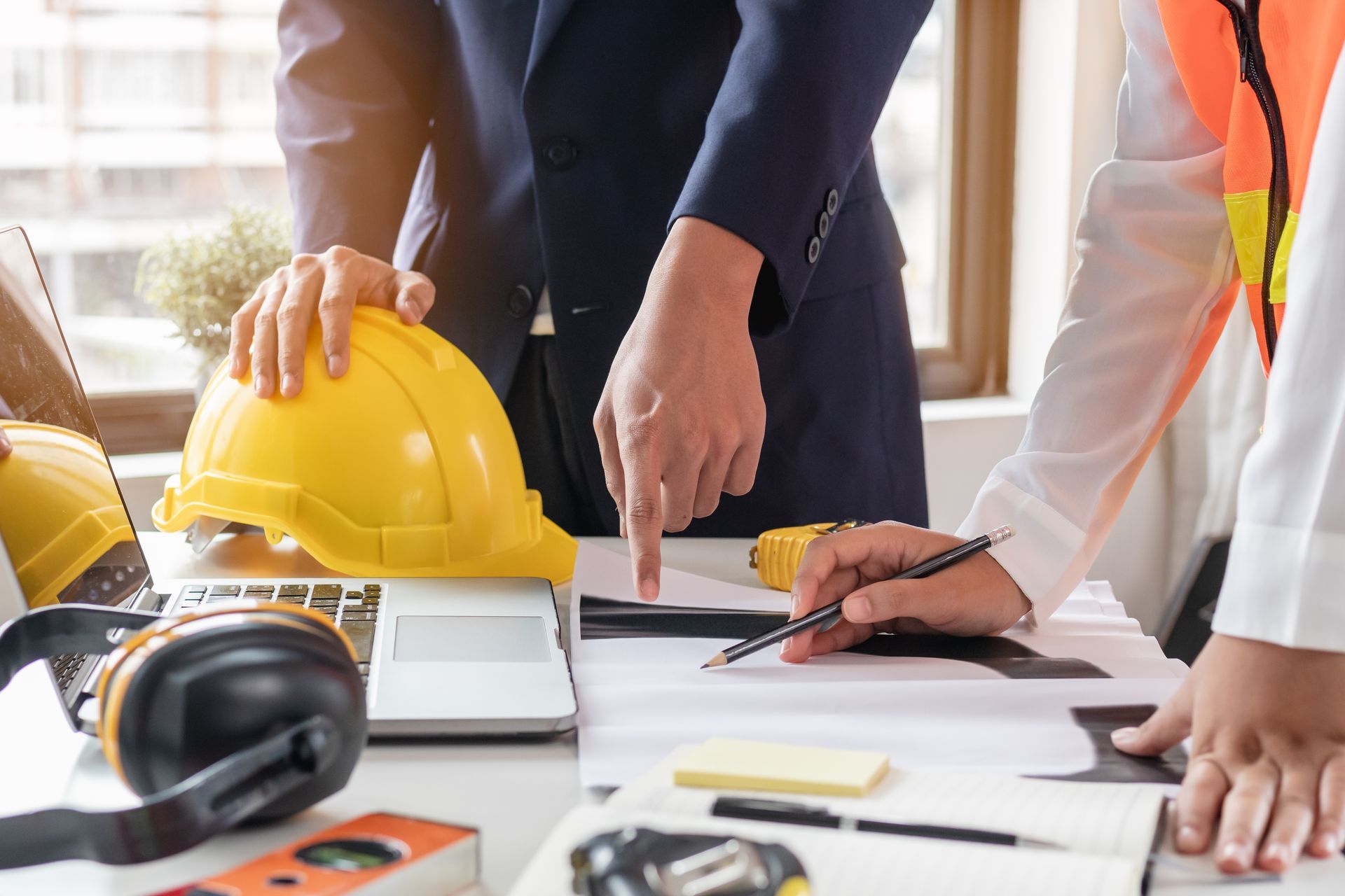 People reviewing construction plans, pointing at a blueprint. Yellow hard hat, laptop, and safety gear on desk.
