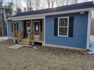 Blue-sided house with a small porch and wooden railings, situated on a grassy area, with trees in the background.