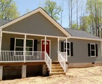 Gray house with a red front door, white porch, and black shutters, set in a wooded area.
