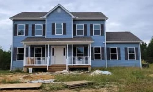 Blue two-story house with white trim, dark shutters, and a front porch, in a grassy field.