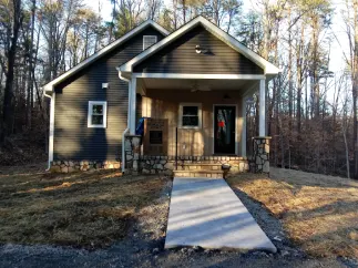 Small, dark-sided house with a porch and stone columns, on a grassy plot. Concrete path leads to front door.