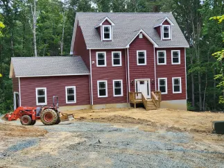 Two-story red house with white trim; tractor parked nearby on dirt lot, surrounded by trees.