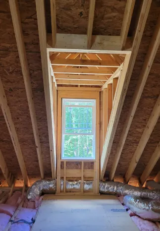 Interior view of attic framing around a window. Wood studs and rafters, insulation, and unfinished plywood.