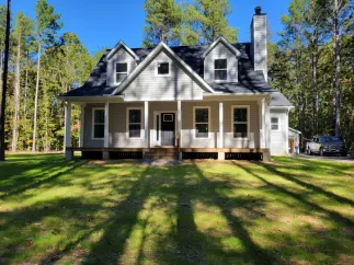 Gray house with a porch, black roof, and chimney, on a green lawn, surrounded by trees.
