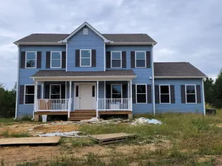 Blue two-story house with white trim, dark shutters, and a porch. Cloudy sky and grassy lot.