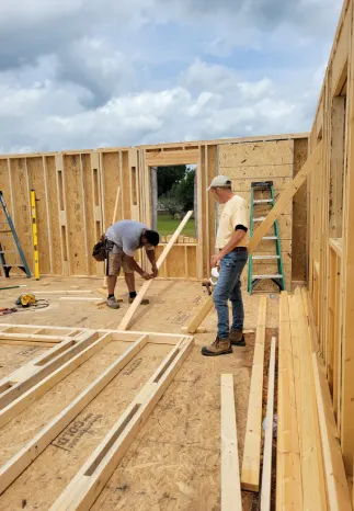 Two construction workers building a wooden frame house. One measures wood, the other observes. Outdoors, cloudy sky.