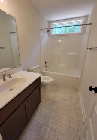 Bathroom with white tub, toilet, and vanity. Brown cabinets, square tile floor, and small window.