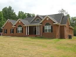 Red brick ranch-style house with black roof, white columns, and large front yard on a sunny day.
