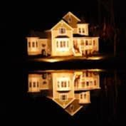 Lit house at night, reflected in still water. Yellow lights glow in the windows against dark background.