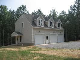 Three-car garage with dormers, light siding, and an attached awning. Set in a wooded area with gravel driveway.