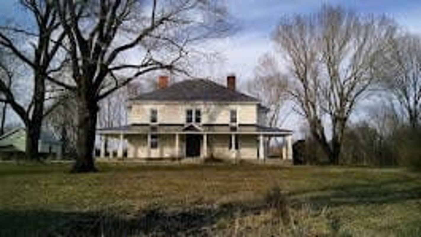 Old, two-story house with wrap-around porch, set in a grassy field; trees line the property.