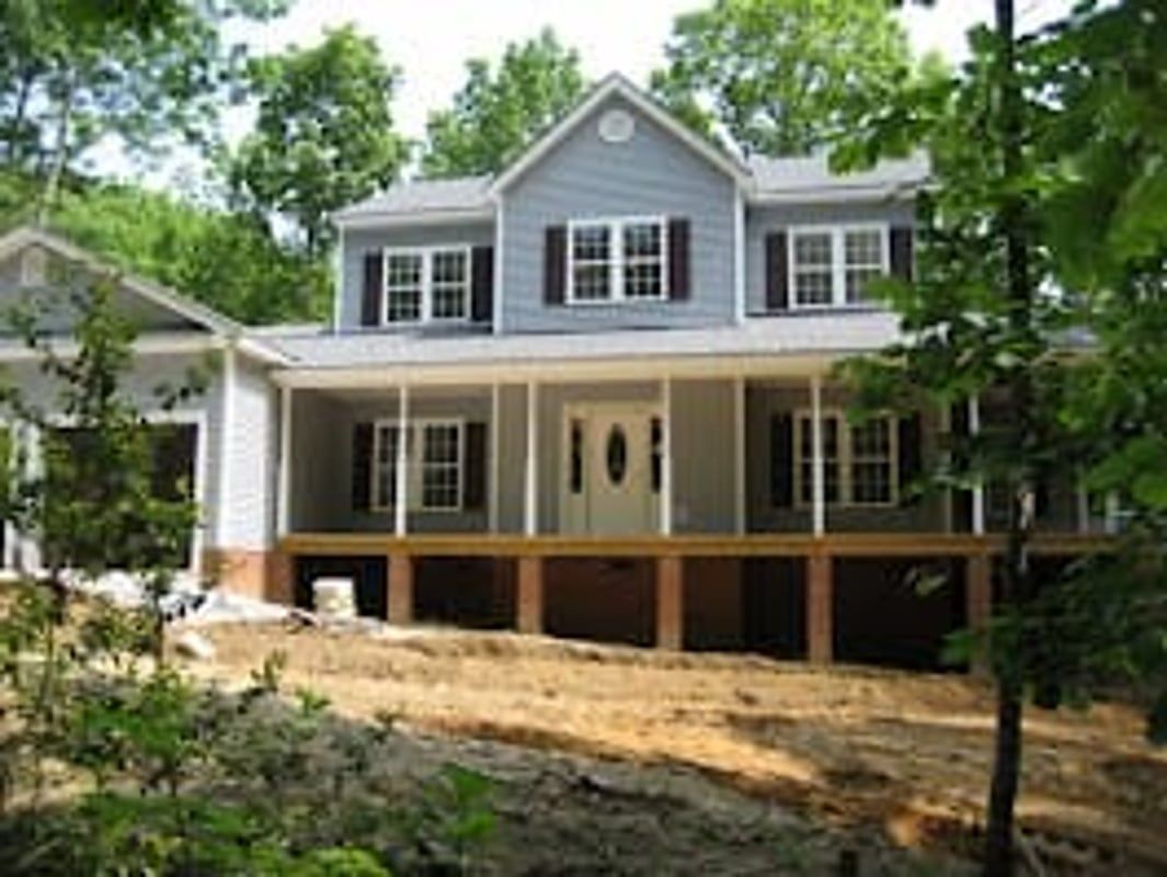Two-story house with a large front porch and blue siding, nestled in a wooded area.