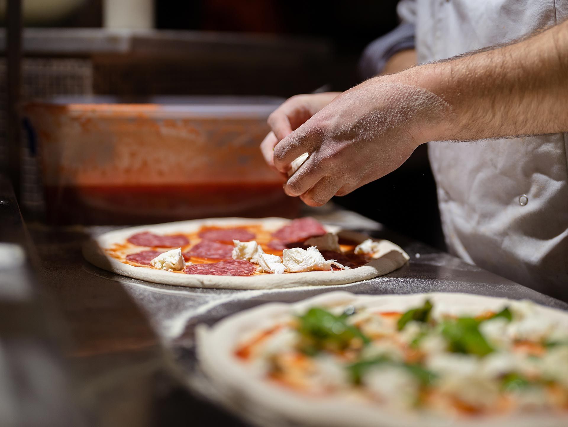 Close-up of a chef’s hands as he adds cheese to a pizza with pepperoni, inside a kitchen. Close-up of a chef’s hands as he adds cheese to a pizza with pepperoni, inside a kitchen.