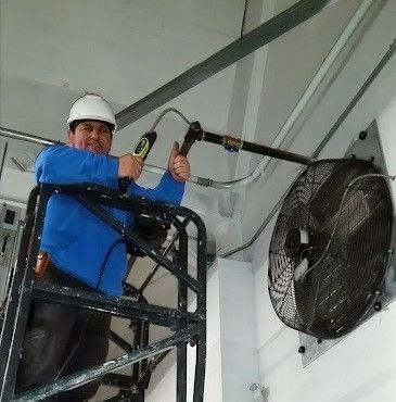 A man is standing on a ladder holding a tape measure in front of a fan.