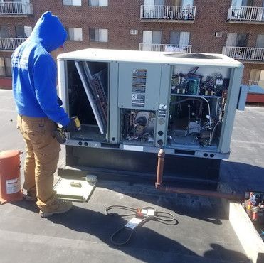A man in a blue hoodie is working on an air conditioner on the roof of a building.