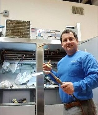 A man in a blue shirt is holding a welding torch in front of a refrigerator.