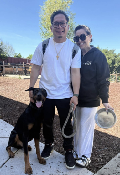 A person with a dog stands beside another person holding a bowl on a paved path in a sunlit outdoor area.